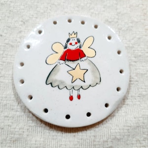 May include: White ceramic round dish with holes around the edge. A hand-painted fairy with a crown, red dress, and yellow wings holds a yellow star.