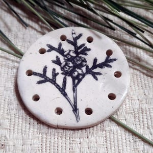 Può includere: Una perlina rotonda in ceramica bianco sporco con un disegno botanico nero disegnato a mano. Il disegno presenta un'illustrazione dettagliata di una pianta con bacche. La perlina ha diversi piccoli fori sul perimetro, per la creazione di gioielli.