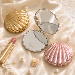 May include: A gold shell-shaped compact mirror, a pink shell-shaped compact mirror, and an open silver-toned compact mirror with the name Elodie engraved on the glass. A lipstick tube and several small white pearls are arranged around the mirrors.