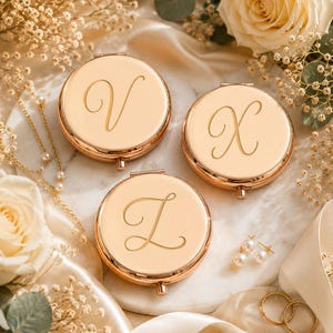 May include: Three round rose gold compact mirrors, each featuring a single engraved cursive letter V, X, or Z on the lid. The metallic finish is polished and reflective, with a small hinge and clasp visible at the bottom of each circular case.