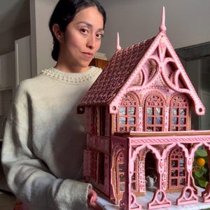 May include: A large, intricate gingerbread house featuring a pink and brown color scheme with detailed icing piping, arched windows, a covered porch, and a peaked roof with decorative finials, constructed with edible materials and structural gingerbread.