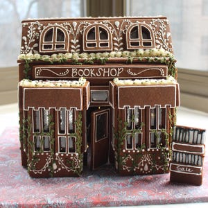 Op de afbeelding: Een peperkoekhuisje in de vorm van een boekwinkel met bruine muren, witte glazuurdetails en groene glazuurranken. De voorkant heeft een bord met de tekst BOOKSHOP en een klein bordje rechts met het woord Sale. Het heeft meerdere ramen en een deur.