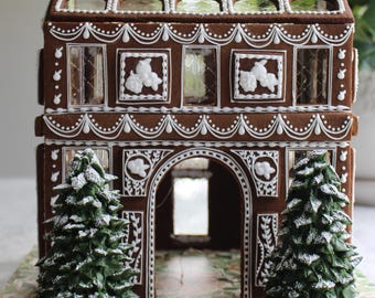 Op de afbeelding: Een peperkoekhuis met bruine muren, witte glazuurversieringen, florale reliëfdetails en doorzichtige vensters. Twee donkergroene, met poedersuiker bestoven dennenbomen van glazuur staan voor de ingang van het gebouw.