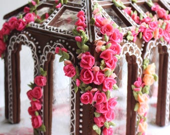 May include: A gingerbread gazebo structure featuring dark brown cookie walls decorated with intricate white royal icing piping. The structure is adorned with numerous small, vibrant pink and peach-colored sugar roses and green icing leaves.