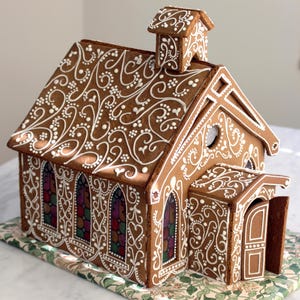 Può includere: Una chiesa di pan di zenzero con intricati dettagli di glassa bianca. La struttura marrone presenta finestre ad arco con vetrate colorate, un piccolo campanile e una porta aperta. Poggia su un tessuto fantasia.