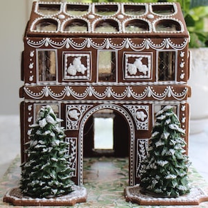 May include: A decorative gingerbread house with brown walls and white icing details. The house has arched windows, decorative trim, and two small green Christmas trees at the front. The roof has arched windows and the house sits on a patterned base.