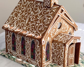 May include: A gingerbread house shaped like a church with brown walls decorated with intricate white icing swirls and dots. The structure features stained glass windows in red, green, and purple, a small steeple, and an open front door.