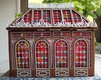 May include: A gingerbread structure designed as a conservatory with brown walls and white icing details. The windows feature translucent red and multicolored panels with a grid pattern. The roof has a white icing trim along the top edge.