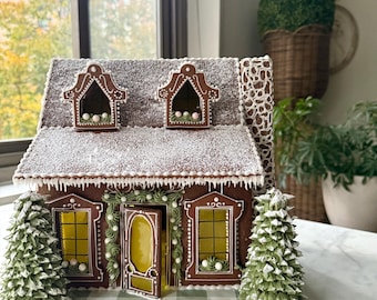 Può includere: Una dettagliata casa di pan di zenzero con un tetto marrone spolverato di zucchero bianco. La casa presenta due abbaini, una porta d'ingresso e due finestre con vetri gialli. Due alberi verdi coperti di neve fiancheggiano la casa.