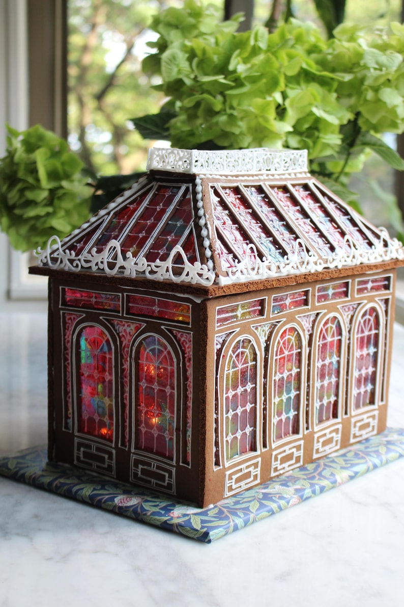 Op de afbeelding: Een decoratief peperkoekhuisje met een glas-in-lood effect. Het dak en de ramen zijn rood en wit, met gedetailleerde witte glazuurdetails. Het staat op een blauw en groen oppervlak met patronen. Groen gebladerte is zichtbaar op de achtergrond.