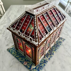Op de afbeelding: Een peperkoekhuisje met glas-in-loodramen en een witte sierlijst. Het dak en de zijkanten hebben rode en oranje glas-in-loodpanelen. Het huis staat op een blauwe basis met bloemmotief.