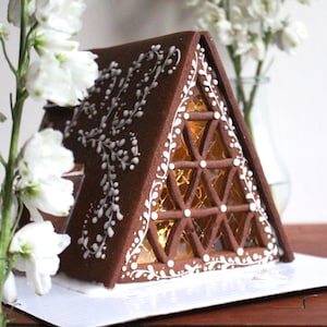 Puede incluir: Una casa de jengibre con un tejado triangular y una fachada con ventanas. La casa está decorada con glaseado blanco con un diseño floral. La ventana está dividida en secciones triangulares. Flores blancas en el fondo.