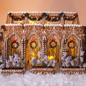 May include: A gingerbread house structure featuring four arched windows with small wreaths, white icing details, and green garlands with red berries. The building is illuminated from within and flanked by two green trees with purple flower boxes.