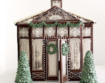 May include: A gingerbread house shaped like a greenhouse with dark brown walls, white icing details, and clear panels. It features a central door with a green icing wreath, green icing garlands, and two small green icing pine trees on each side.
