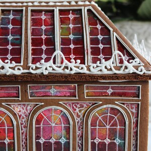 Op de afbeelding: Een peperkoekhuisje met glas-in-loodramen. Het huis heeft een bruine buitenkant met witte glazuurdetails en levendige rode, roze en gele glas-in-loodramen. Het dak heeft een decoratieve witte glazuurrand.