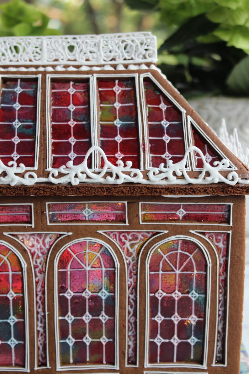 Op de afbeelding: Een peperkoekhuisje met glas-in-loodramen. Het huis heeft een bruine buitenkant met witte glazuurdetails en levendige rode, roze en gele glas-in-loodramen. Het dak heeft een decoratieve witte glazuurrand.