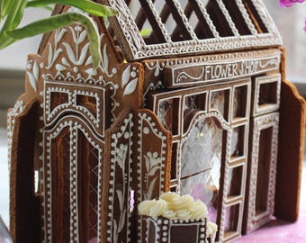 May include: A gingerbread house shaped like a flower shop with brown walls and white icing details. The structure features a gabled roof, arched windows, and a sign reading FLOWER SHOP. A small rectangular planter with white icing flowers sits in front.