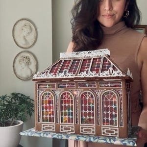 Op de afbeelding: Een gedetailleerd peperkoekhuis met boogvensters en kleurrijke glas-in-lood effect panelen. Het dak heeft een decoratieve witte rand. Het huis staat op een patroon, wat wijst op een feestelijk thema, mogelijk voor Kerstmis.