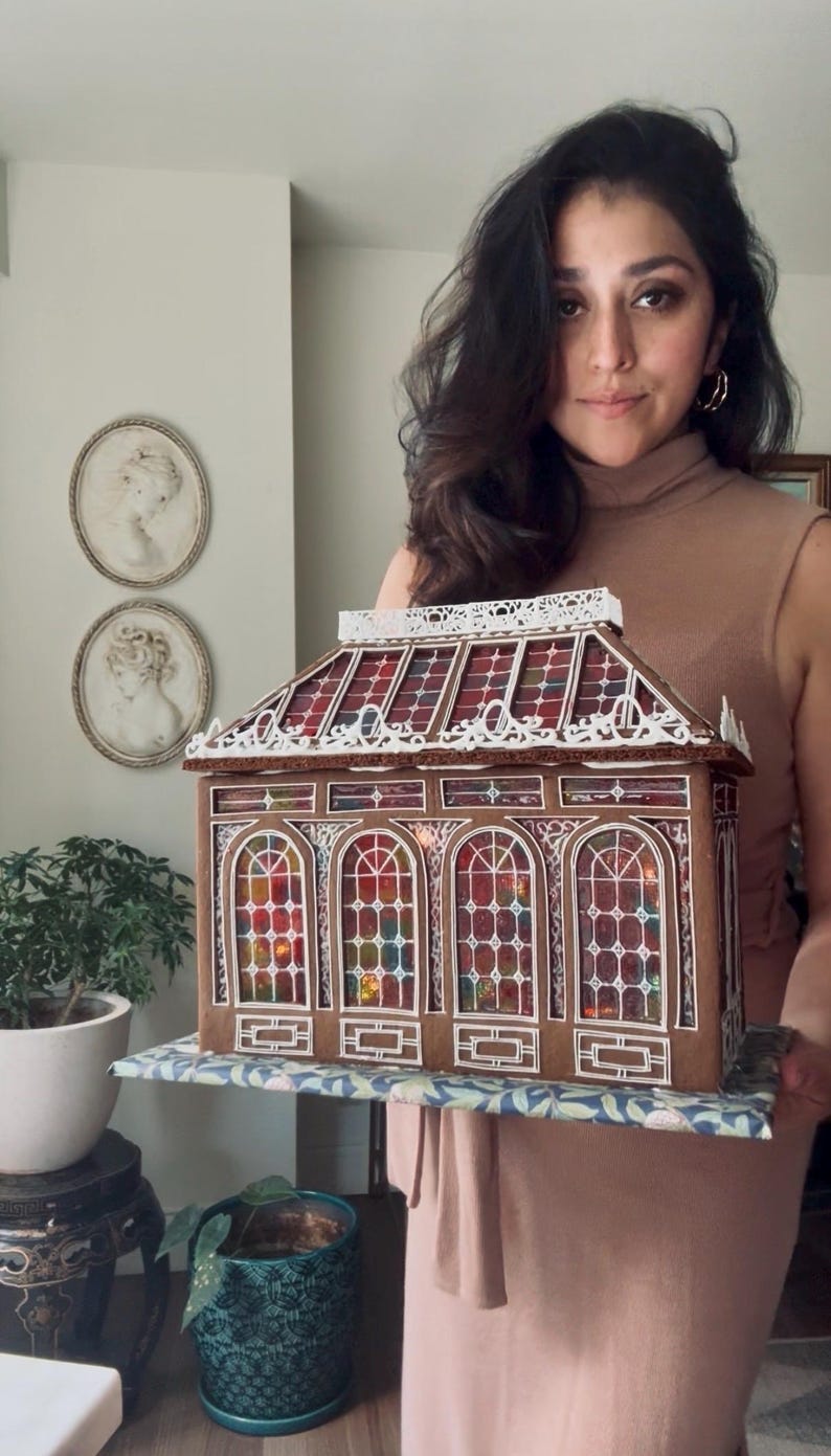 Op de afbeelding: Een gedetailleerd peperkoekhuis met boogvensters en kleurrijke glas-in-lood effect panelen. Het dak heeft een decoratieve witte rand. Het huis staat op een patroon, wat wijst op een feestelijk thema, mogelijk voor Kerstmis.