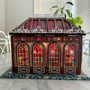 Op de afbeelding: Een peperkoekhuisje met een glas-in-loodraamontwerp. Het dak en de ramen zijn rood, oranje en blauw. Het huis heeft witte decoratieve randen en staat op een blauw oppervlak met patronen.
