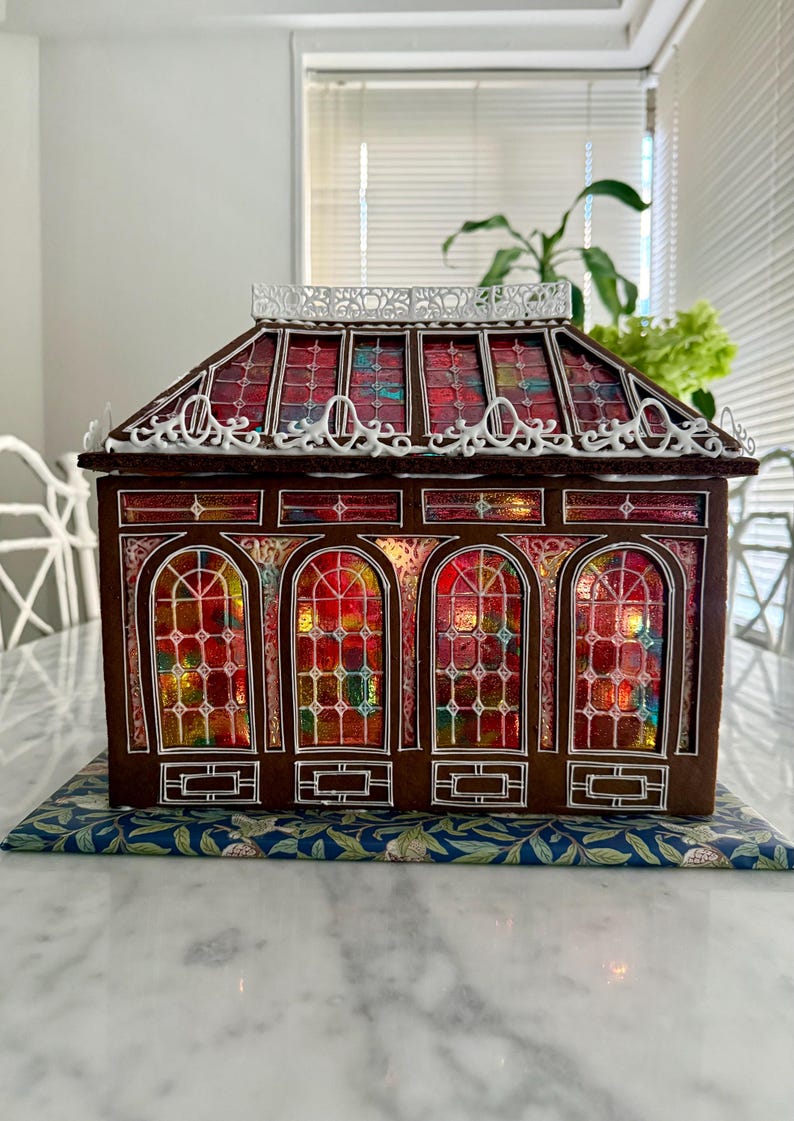 Op de afbeelding: Een peperkoekhuisje met een glas-in-loodraamontwerp. Het dak en de ramen zijn rood, oranje en blauw. Het huis heeft witte decoratieve randen en staat op een blauw oppervlak met patronen.