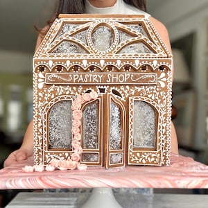 May include: A gingerbread house shaped like a pastry shop with white icing scrollwork, clear windows with glittery accents, and a vertical vine of small pink icing roses. The structure sits on a pink and white marbled base on a white pedestal stand.