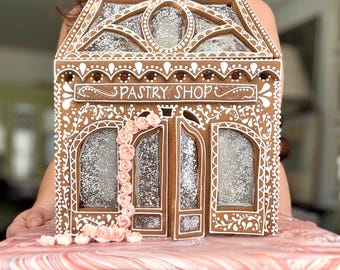 Pode incluir: Uma casa de gengibre em forma de pastelaria com detalhes em glacê branco, janelas transparentes com brilhos e uma trepadeira vertical de pequenas rosas de glacê rosa sobre uma base marmorizada rosa e branca num suporte branco.