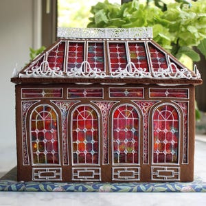 Op de afbeelding: Een peperkoekhuisje met een bruine gevel en een rood-geel glas-in-lood raamontwerp. Het dak heeft een decoratieve witte rand. Het huis staat op een blauw bloemenpatroon. Het thema is vakantiedecoratie.