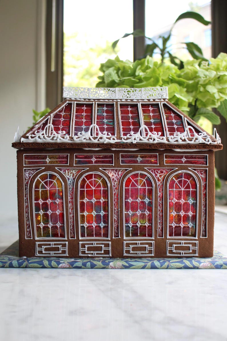 Op de afbeelding: Een peperkoekhuisje met een bruine gevel en een rood-geel glas-in-lood raamontwerp. Het dak heeft een decoratieve witte rand. Het huis staat op een blauw bloemenpatroon. Het thema is vakantiedecoratie.