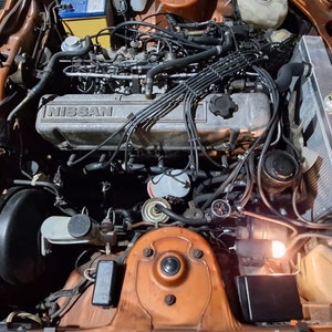 May include: Close-up view of a car engine bay with a brown and black engine. The engine has a silver valve cover with the word "NISSAN" printed on it. The engine is surrounded by various hoses and wires.