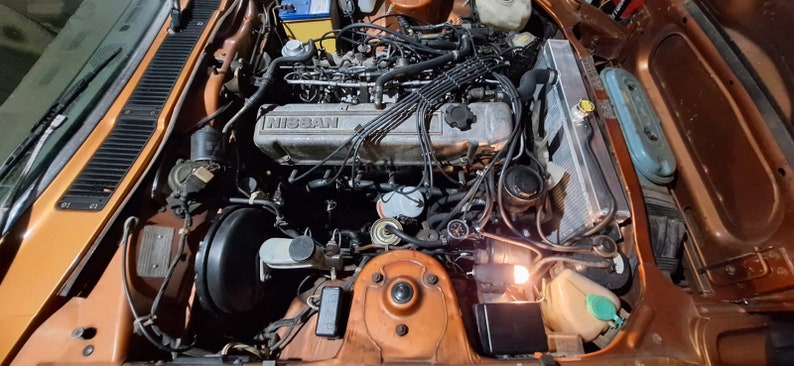 May include: Close-up view of a car engine bay with a brown and black engine. The engine has a silver valve cover with the word "NISSAN" printed on it. The engine is surrounded by various hoses and wires.