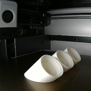 Puede incluir: Una impresora 3D con tres objetos blancos en forma de cono en primer plano. La impresora es blanca y negra, con el texto "Online - Idle" en la pantalla. Los objetos están dispuestos en fila.