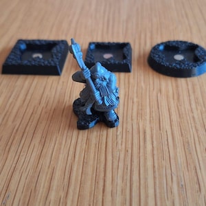 May include: A miniature warrior figurine with a sword, next to three black bases. The bases are square and round, with textured edges. The warrior is dark grey, and the bases are black.