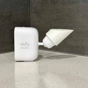 May include: A white security camera with the brand name "eufy SECURITY" printed on the front. The camera has a rectangular body and a cone-shaped extension. It is mounted on a light-coloured surface.