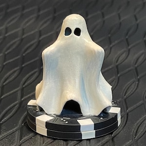 May include: A white ghost figurine with black eyes, sitting on a black and white poker chip.