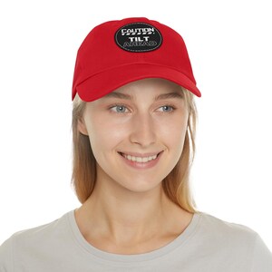 Puede incluir: Gorra de béisbol roja brillante con visera curva. La gorra presenta un parche negro con las palabras "CAUTION" encima de "TILT AHEAD" en texto blanco. El sombrero está hecho de un material suave.