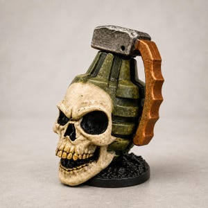 May include: A decorative figurine shaped like a human skull integrated with a green, segmented hand grenade body. It features a metallic silver top lever and a textured copper-colored handle, all resting on a circular black base.