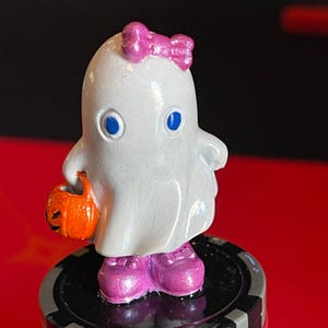 May include: A white ghost figurine with a pink bow, blue eyes, and pink shoes, holding an orange pumpkin-shaped treat bag. The figurine is standing on a stack of poker chips, with a red and black background.