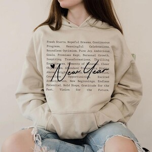 Op de afbeelding: Een beige hoodie met een zwarte print die luidt: "Fresh Starts. Hopeful Dreams. Continuous Progress. Meaningful Celebrations. Boundless Optimism. Pure Joy. Ambitious Goals. Promises Kept. Personal Growth. Inspiring Transformations. Thrilling Experiences. Moments of Renewal. Clear Objectives. Abundant Prosperity. Rising Above. New Year. Countless Opportunities. Excited Anticipation. New Beginnings. Endless Potential. Bold Steps. Gratitude for the Past. Vision for the Future."