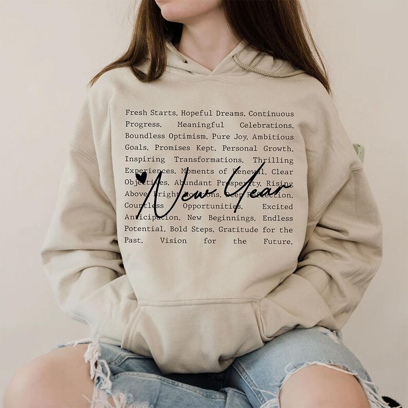 Op de afbeelding: Een beige hoodie met een zwarte print die luidt: "Fresh Starts. Hopeful Dreams. Continuous Progress. Meaningful Celebrations. Boundless Optimism. Pure Joy. Ambitious Goals. Promises Kept. Personal Growth. Inspiring Transformations. Thrilling Experiences. Moments of Renewal. Clear Objectives. Abundant Prosperity. Rising Above. New Year. Countless Opportunities. Excited Anticipation. New Beginnings. Endless Potential. Bold Steps. Gratitude for the Past. Vision for the Future."