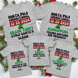 May include: Five gray t-shirts with a Christmas theme. Each shirt has a different design with a North Pole Correctional theme and a funny crime. The text on the shirts reads: "North Pole Correctional 12-25-2024" and then a crime such as "Grand Theft", "Kidnapping", "Arson", "Theft", and "Public Disturbance".