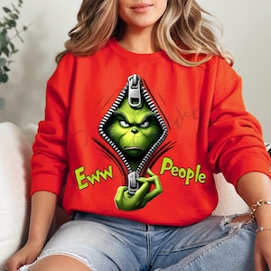 Könnte beinhalten: Roter Pullover mit einem Grafikdesign des grünen Grinch-Gesichts, das aus einem Reißverschluss herausragt. Die Worte "Eww People" sind grün auf dem Pullover gedruckt. Die Hand des Grinch ist ebenfalls sichtbar.