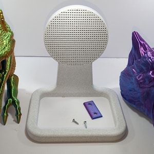 May include: A white 3D printed stand with a round speaker grille, a small metal plate, and two screws. The stand is on a white surface.