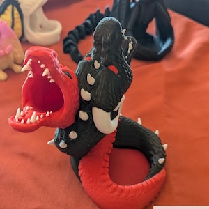 May include: A 3D-printed, multicolor dragon dice tower and roller. The dragon is black and red with white accents and sharp teeth. The text "Dueling Dragon Dice Tower and Roller Multicolor!" is at the top.