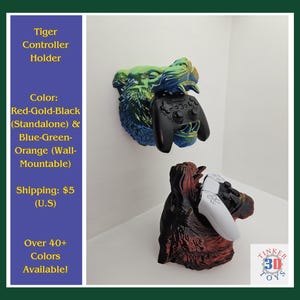 May include: Two 3D printed tiger head controller holders. One is blue, green, and gold with a black controller. The other is red and black with a white controller. The text "TINKER 3D TOYS" is visible in the bottom right corner.