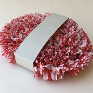 May include: A red and white paper pom-pom wrapped in a white paper band. The pom-pom is made of thin strips of paper that are curled and twisted to create a fluffy texture.