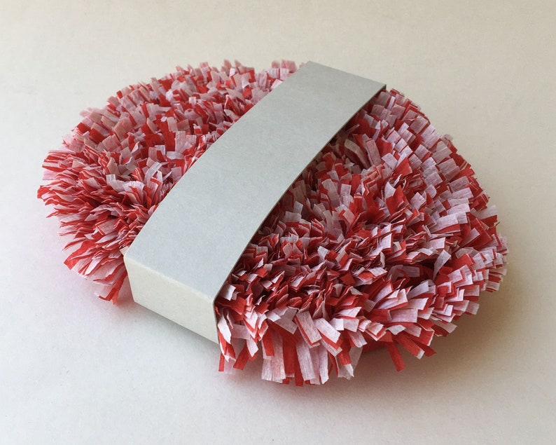 May include: A red and white paper pom-pom wrapped in a white paper band. The pom-pom is made of thin strips of paper that are curled and twisted to create a fluffy texture.