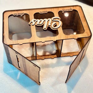 May include: A rectangular wooden money box with three clear acrylic compartments labelled share, save, and spend. The name Silas is cut out in wood and attached to the top. The box features a light brown wood grain finish and a hinged front panel.