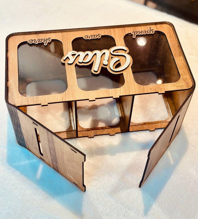 May include: A rectangular wooden money box with three clear acrylic compartments labelled share, save, and spend. The name Silas is cut out in wood and attached to the top. The box features a light brown wood grain finish and a hinged front panel.