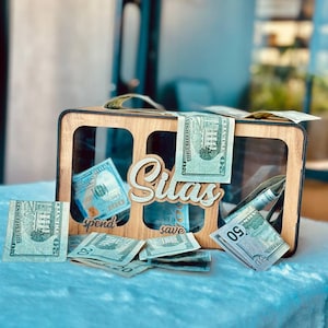 May include: A rectangular wooden money box with three clear acrylic windows and the name Silas in script lettering. The box features labels for spend and save, and is filled with various United States paper currency bills including twenties and fifties.